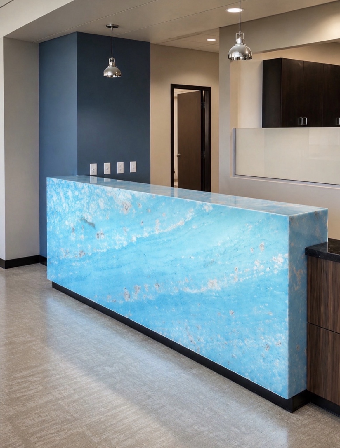 Illuminated blue onyx reception counter with a matching top and full-height front, surrounded by dark cabinetry and pendant lights