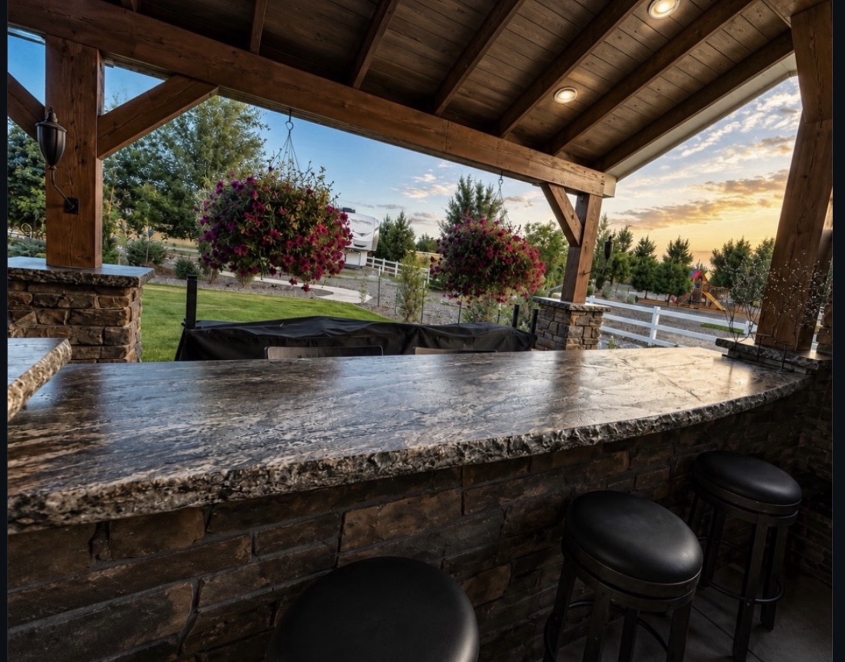 Covered outdoor bar with a patterned stone countertop and textured edge, stone base, black stools and timber beams overlooking a garden at sunset