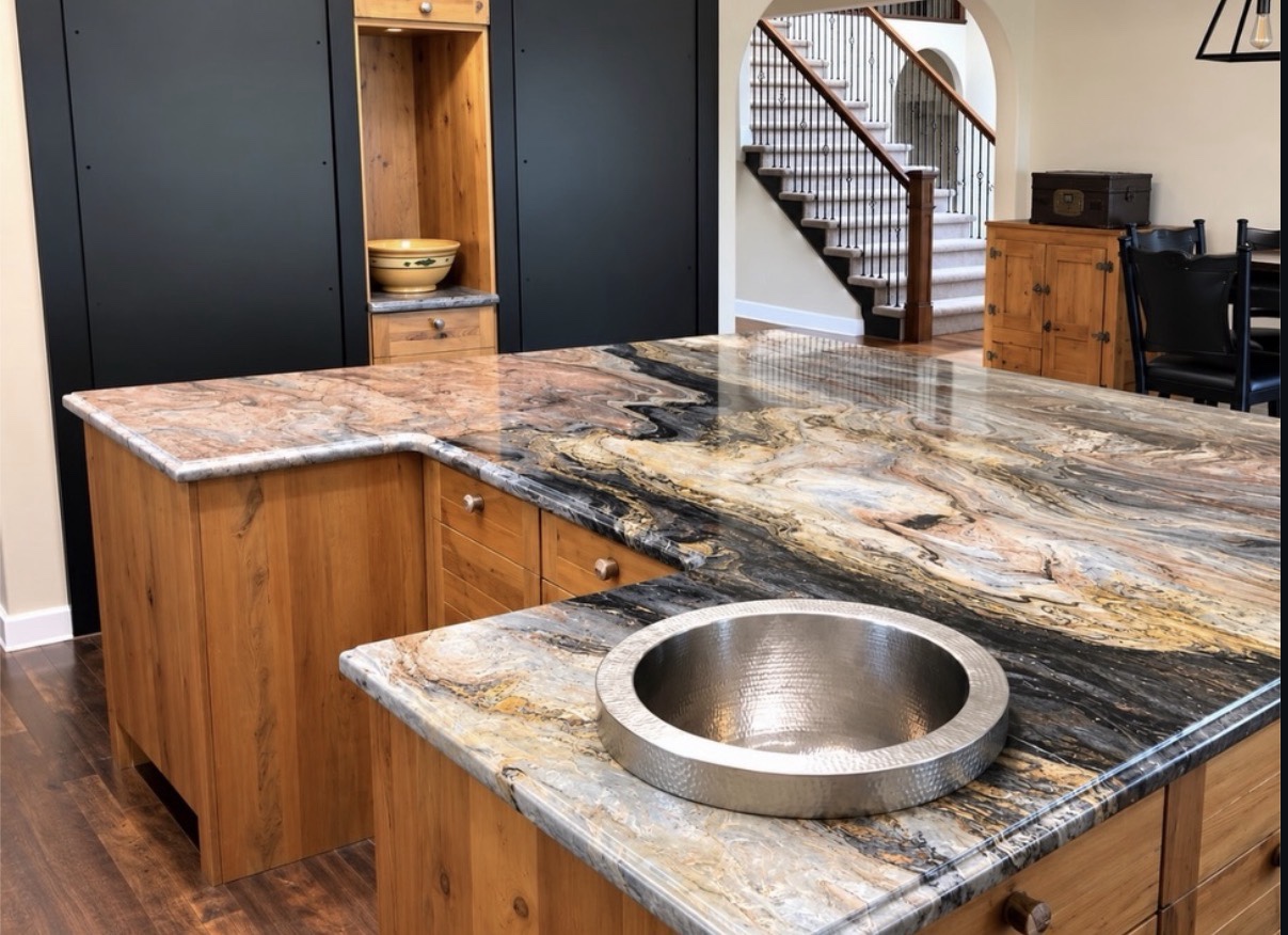 Polished stone countertop with flowing black, gold, cream and pink patterns above wood cabinetry, with a round hammered metal sink