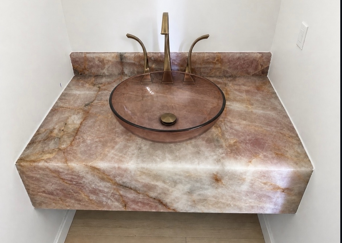 Floating quartzite vanity top with a deep mitered apron, matching backsplash, round vessel sink and brass faucet