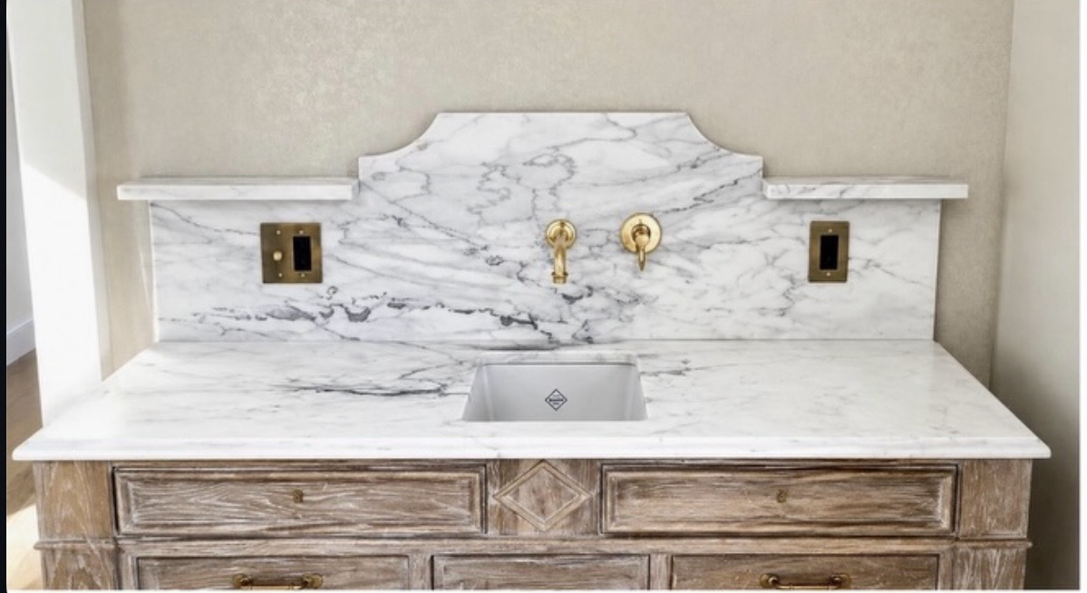 White marble vanity countertop with gray veining, a shaped marble backsplash, undermount sink and wall-mounted brass fixtures above a wood cabinet
