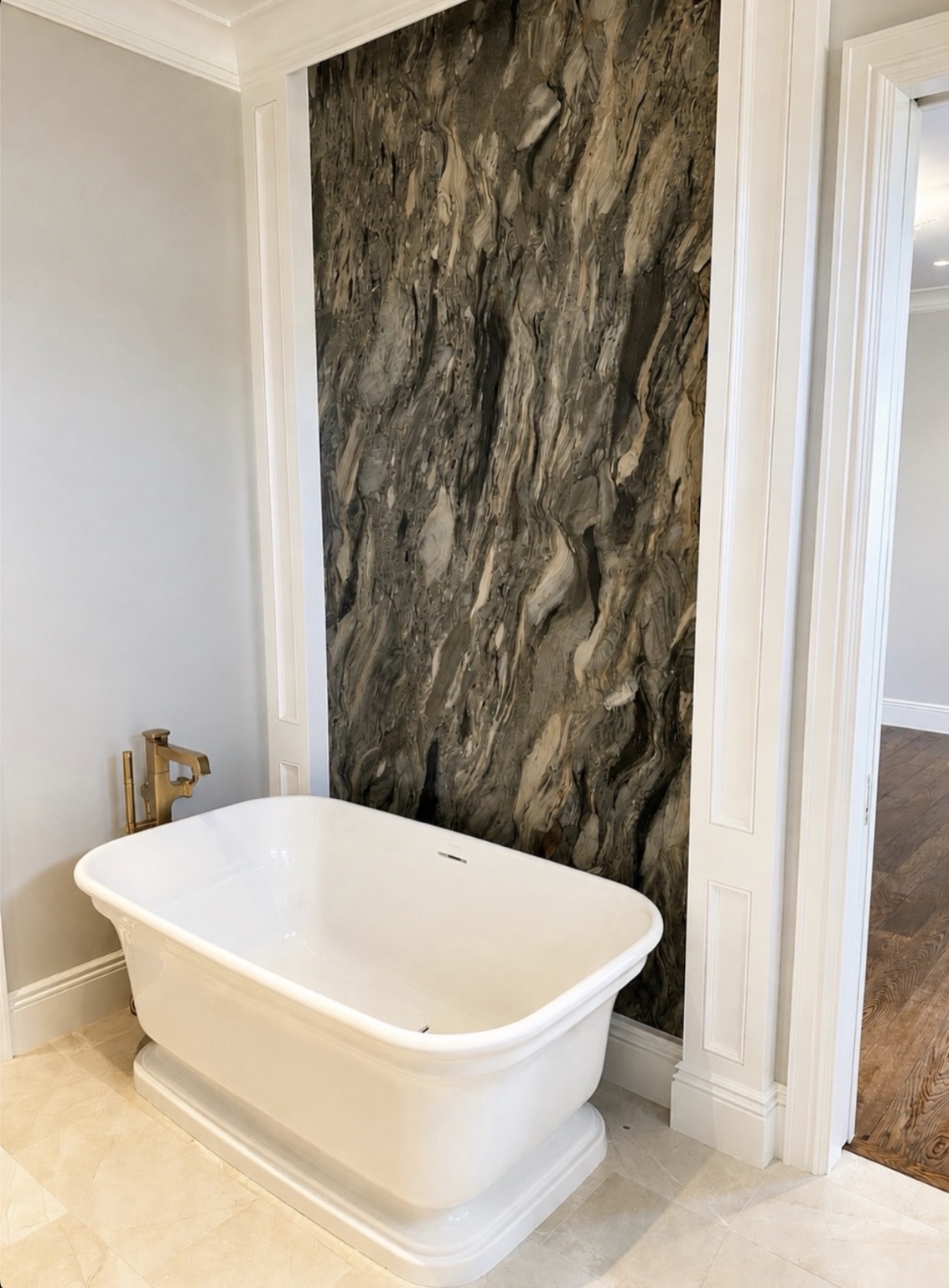 Freestanding white bathtub with a brass faucet in front of a tall patterned stone feature wall framed by white trim