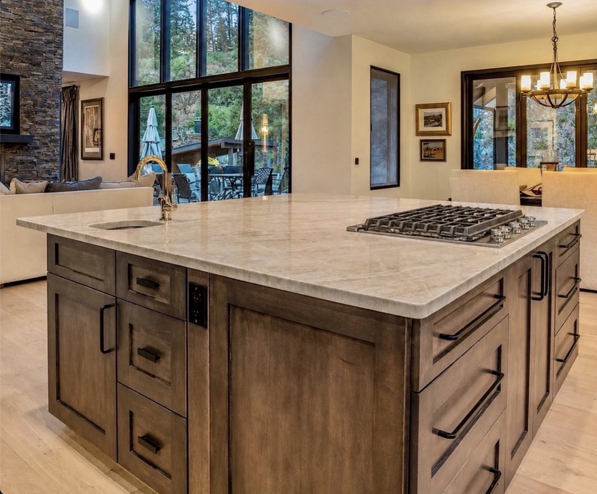 Large pale stone countertop on a wood kitchen island with an undermount sink, brass faucet and gas cooktop overlooking an open living and dining area